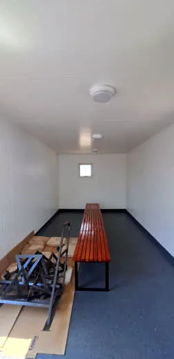 Empty change room with white walls, dark speckled floor and a timber slat bench, with packaging and steel frames on the floor at left.