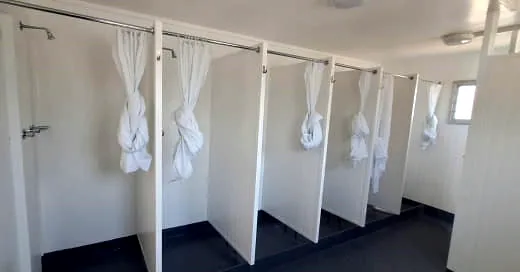 Row of shower stalls with white partitions and tied-back white curtains on a dark floor.