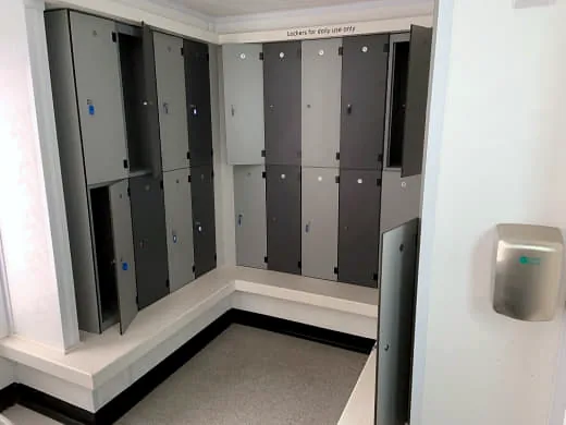 Change room with grey and charcoal lockers in an L-shaped bank above a white bench base, a 'Lockers for daily use only' sign and a wall-mounted hand dryer.