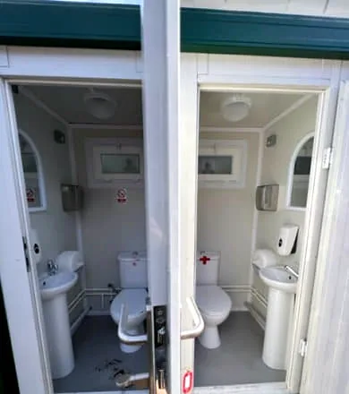 Two toilet cubicles seen through open doors, each with a toilet, pedestal basin, hand towel dispenser and window.