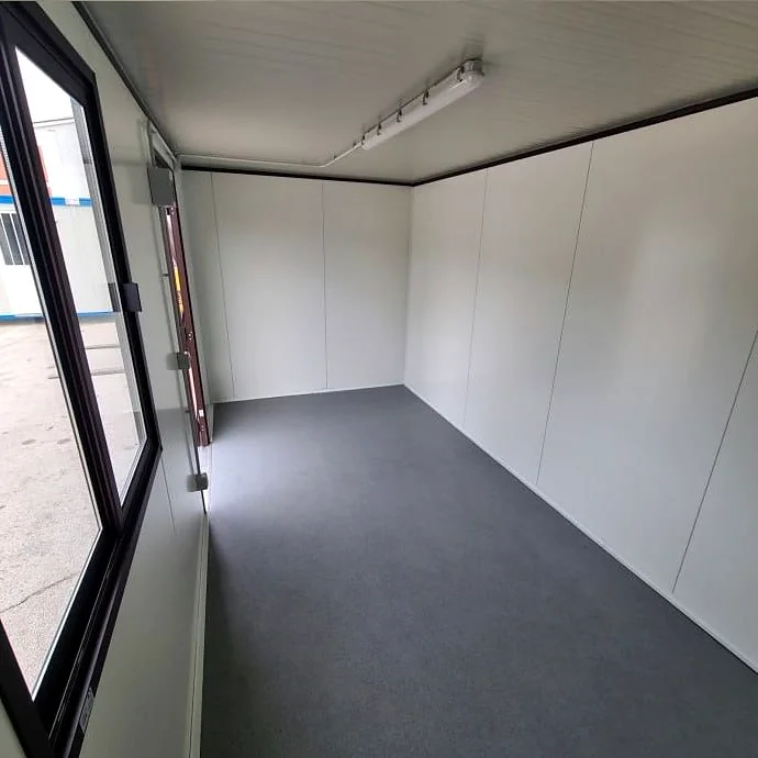 Empty interior of a portable cabin with white wall panels, grey floor covering, a ceiling light and a window beside the door.