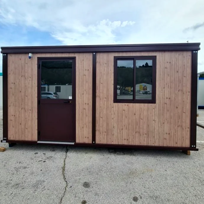 Front view of a portable cabin with timber-look wall panels, dark brown trim, a half-glazed door and a two-pane window, sitting on a concrete yard.