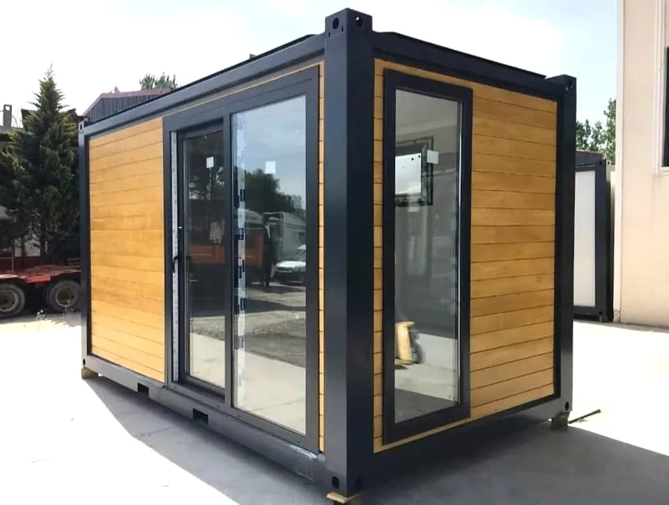 Timber-look cabin with black frame and a large glass front, on a concrete yard.
