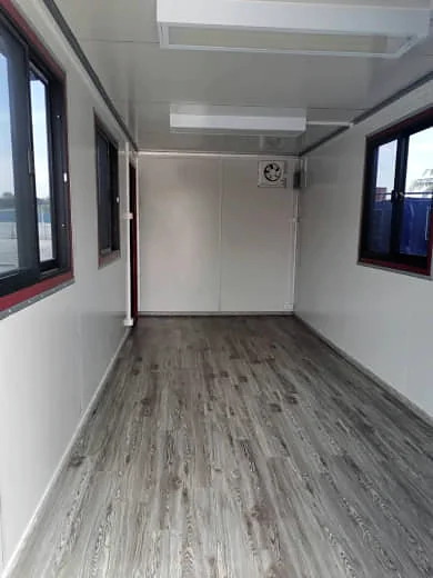 Lined container office interior with white wall panels, timber-look flooring, ceiling lights, an exhaust fan and windows on both sides.