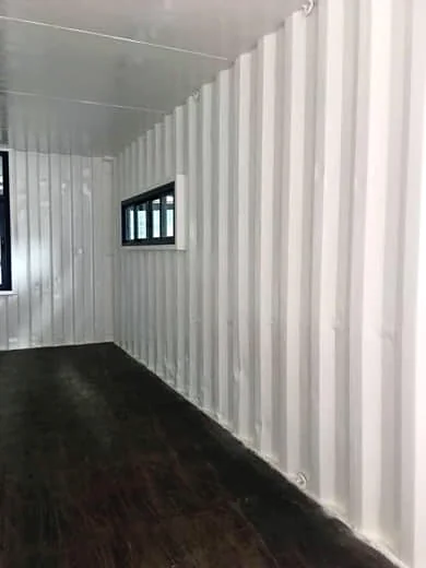 Interior of a shipping container with white-painted corrugated steel walls, a dark timber floor and a small horizontal window.