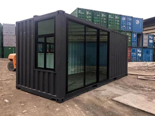 Black shipping container office with full-height black-framed glass panels and a window on the side, in a container yard.