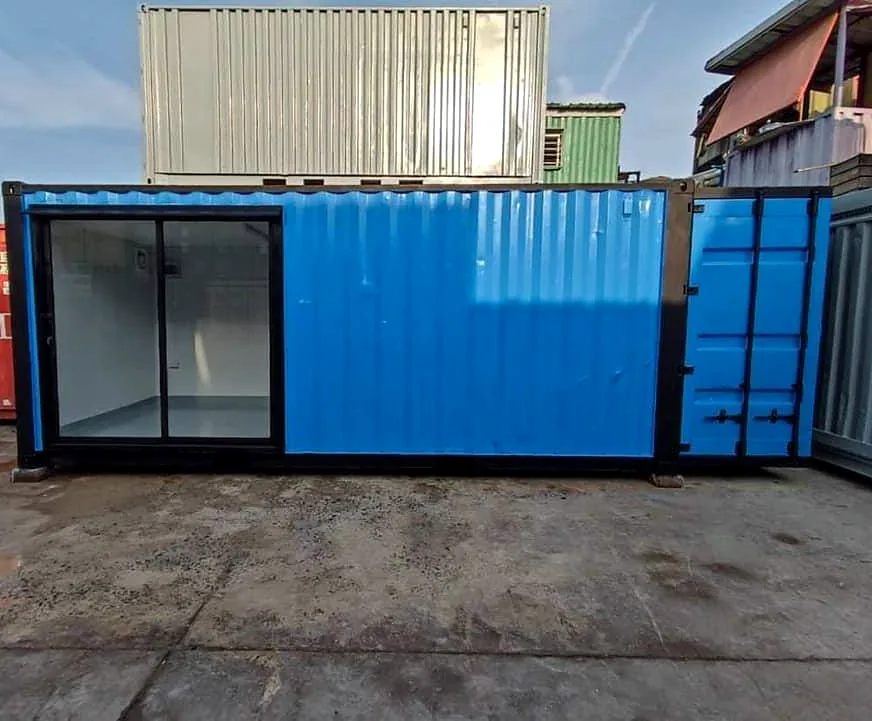 Blue shipping container converted with a large black-framed glass front, on a concrete yard.