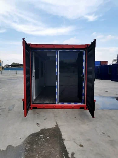 End view of a black and red shipping container with its doors open, showing glass sliding doors behind them.