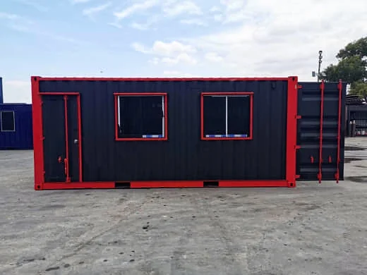 Side view of a black shipping container office with red trim, a personnel door and two windows, with the end doors open.