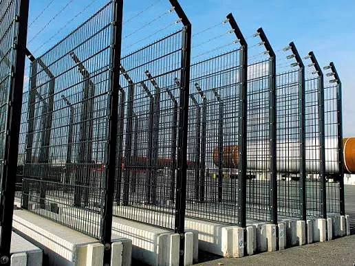 Black welded-mesh security fence panels with angled barbed-wire arms, set on concrete blocks.