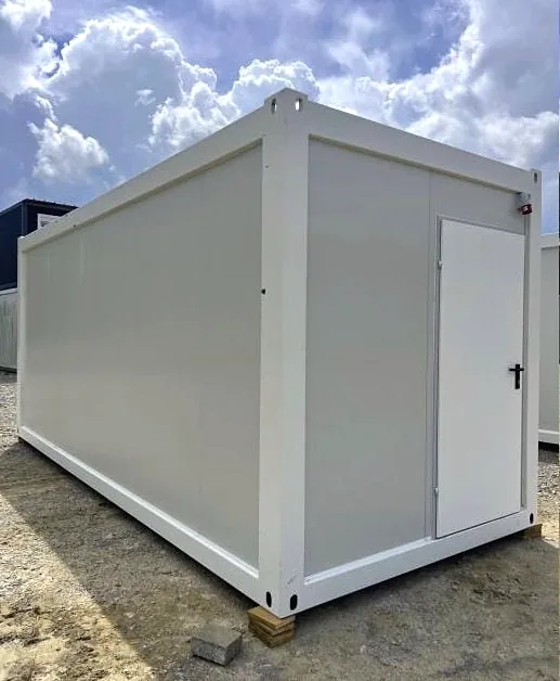 White flatpack container building with a white door on the side, on gravel under a cloudy sky.