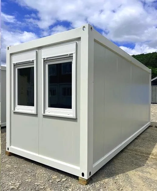 White flatpack container building with two windows on the end, on gravel under a cloudy sky.