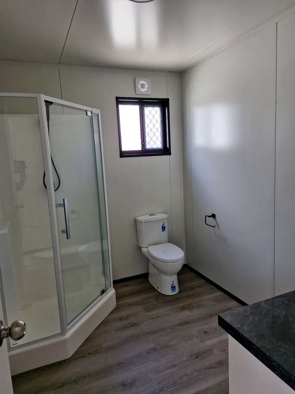 Bathroom with a corner glass shower, toilet, exhaust fan and a security-screened window.