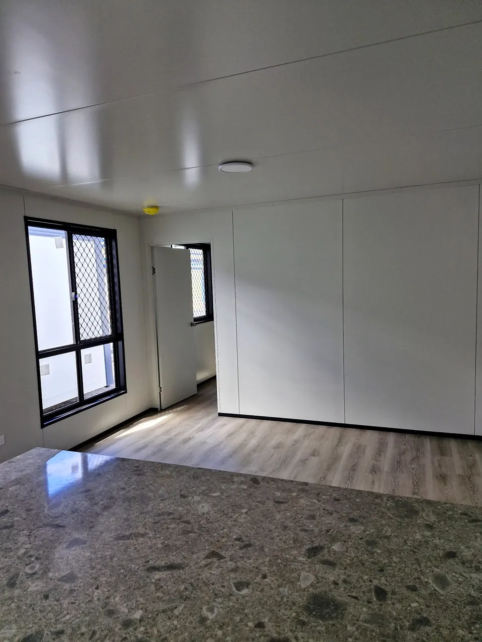 Living area viewed over a stone-look benchtop, with white walls and security-screened windows.