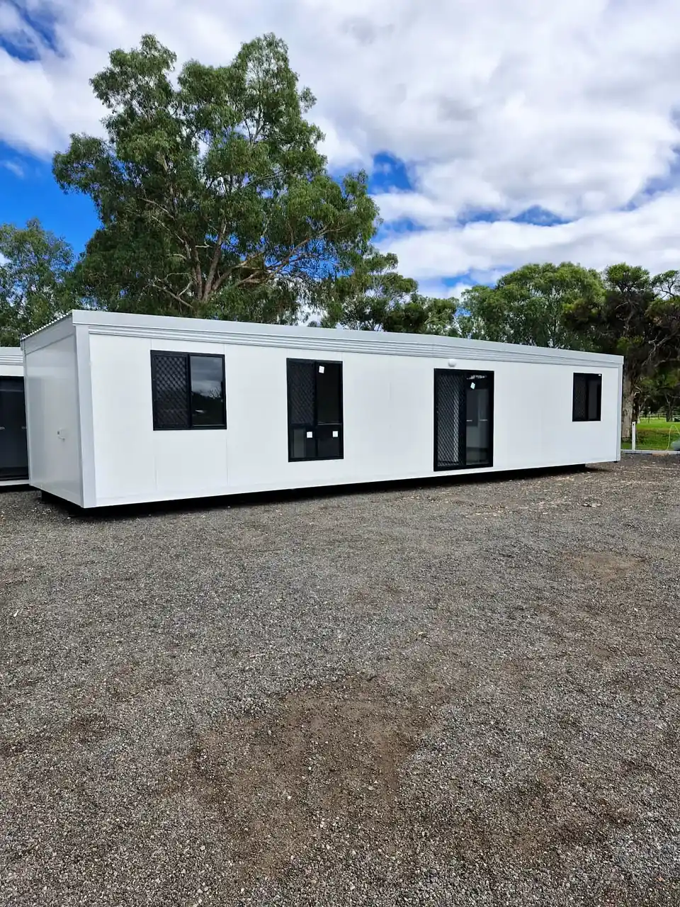Long white portable home with black windows and doors on a gravel site with gum trees behind.