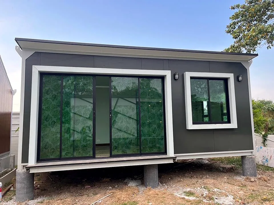 Front of a small charcoal cabin with a wide glass sliding door and window, raised on concrete piers.
