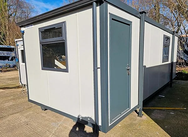 White and grey modular building with a grey door and window, on a paved yard with bare trees behind.