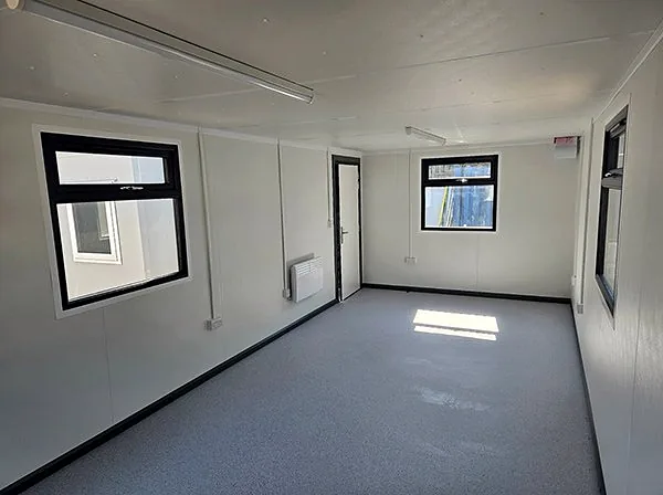 Empty modular building interior with white panel walls, grey speckled floor, black-framed windows and a wall heater.