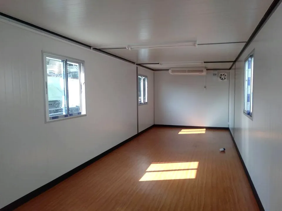 Long empty modular interior with white walls, timber-look floor, sliding windows and a wall-mounted air conditioner and exhaust fan at the far end.