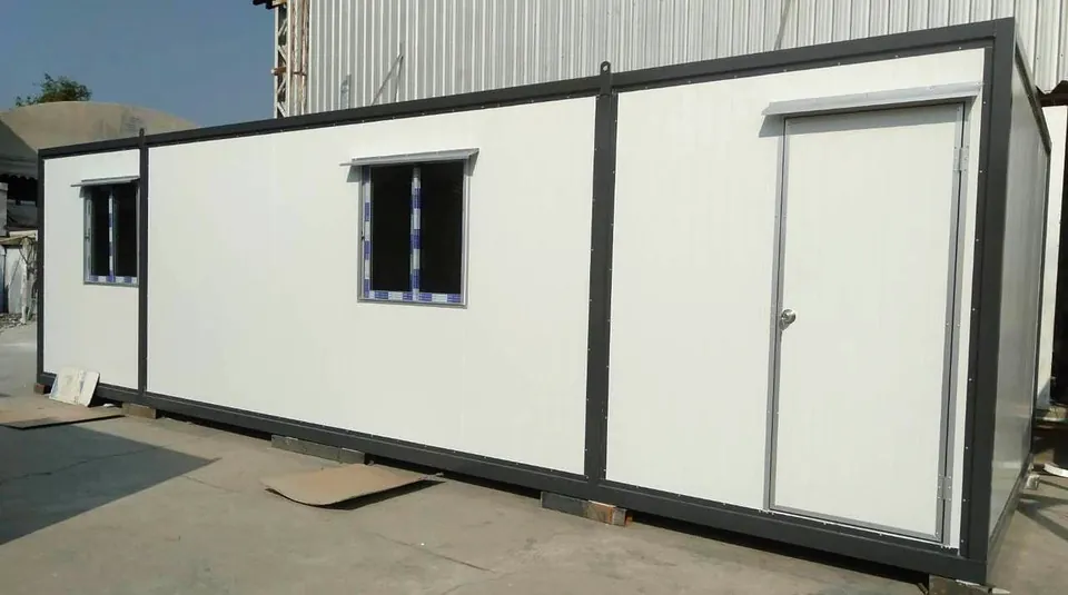 Long white modular building with grey frame, one door and two windows, in front of a factory shed.