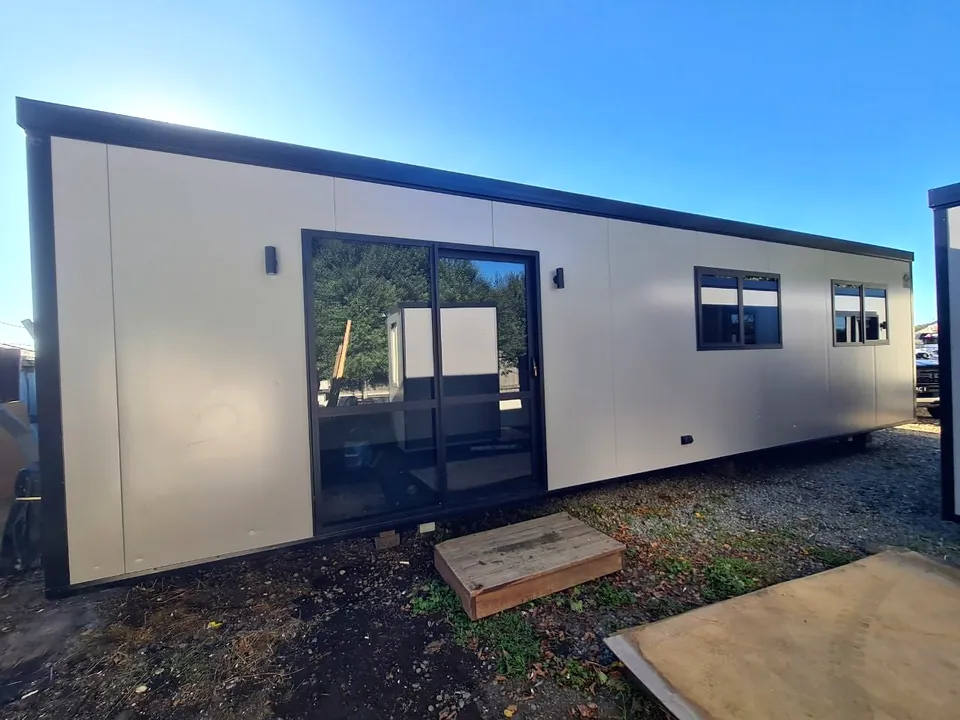 Long cream portable cabin with black trim, a black-framed glass sliding door and two windows, on a gravel site.
