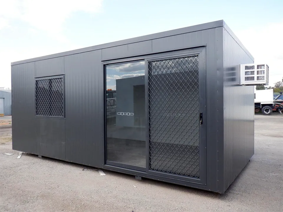 Charcoal portable office with glass sliding door, mesh security screens and a box air conditioner, on a gravel yard.
