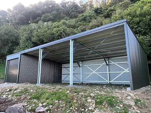 Dark steel open-bay farm shed on a hillside with bush behind.